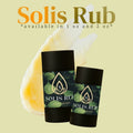 Two black Solis Rub body rub sticks with green leaf-patterned labels, shown against a pale yellow-green background with a translucent yellow wax smear and the text 'Solis Rub' and 'available in 1 oz and 2 oz'.