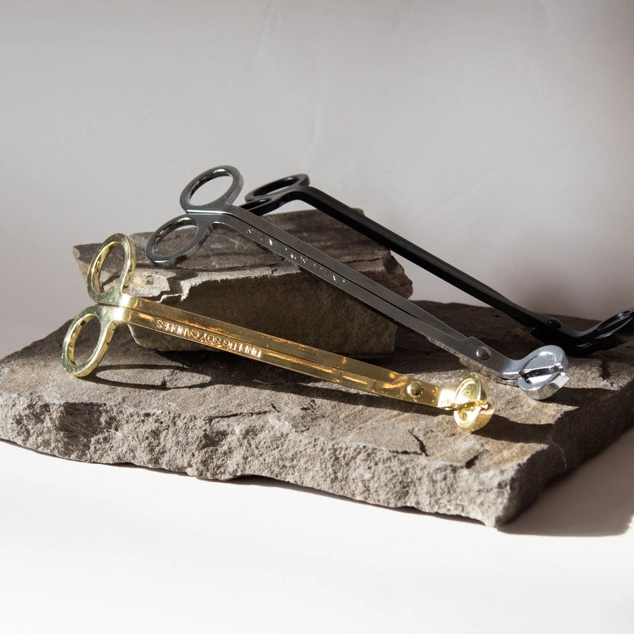 A pair of gold-toned metal forceps with a circular loop at the tip, resting on a textured gray stone slab with a black-handled tool behind it, photographed in a studio setting.